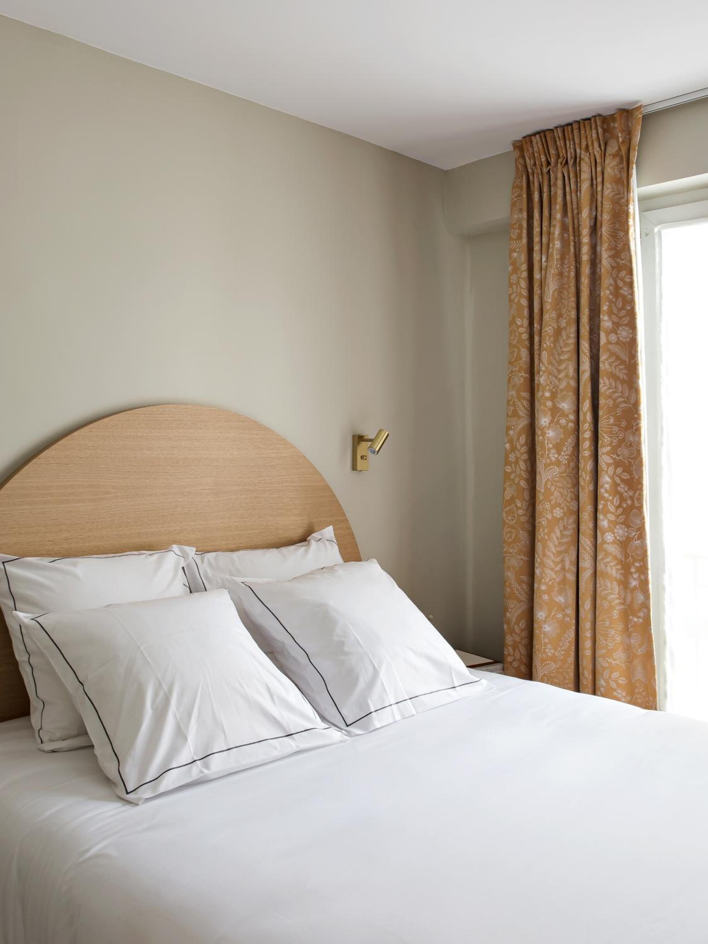 Un lit bien fait, avec du linge de lit blanc et une tête de lit en bois, une lampe fixée au mur et une fenêtre avec des rideaux.
