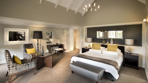 A spacious bedroom with a large bed, a desk, and a chair. There is a desk lamp, a chair, and a bedside table with a lamp.
