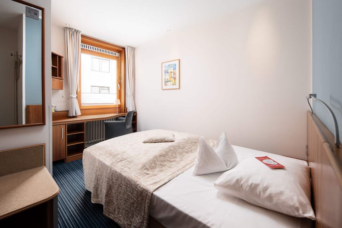 A hotel room with a bed, a desk, a chair, a window with curtains, a radiator, and a framed picture on the wall.