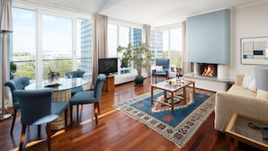 A spacious living room with a large window, a fireplace, and a dining area with a glass table and blue chairs. - Hotel Baseler Hof (Hamburg)