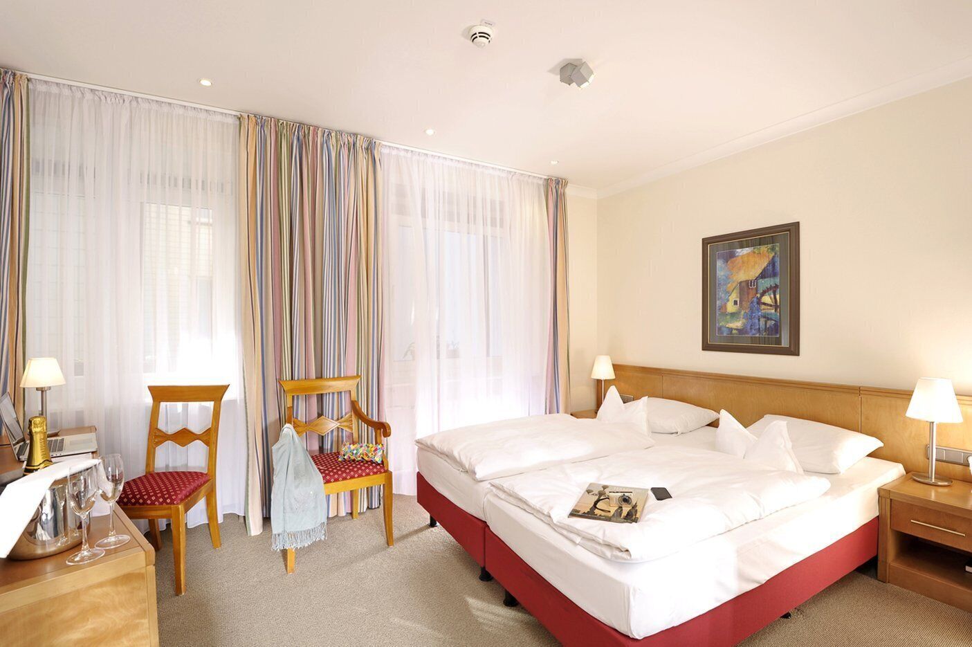 Habitación de hotel con una cama grande, una silla de madera, un escritorio con lámpara y un cuadro en la pared.