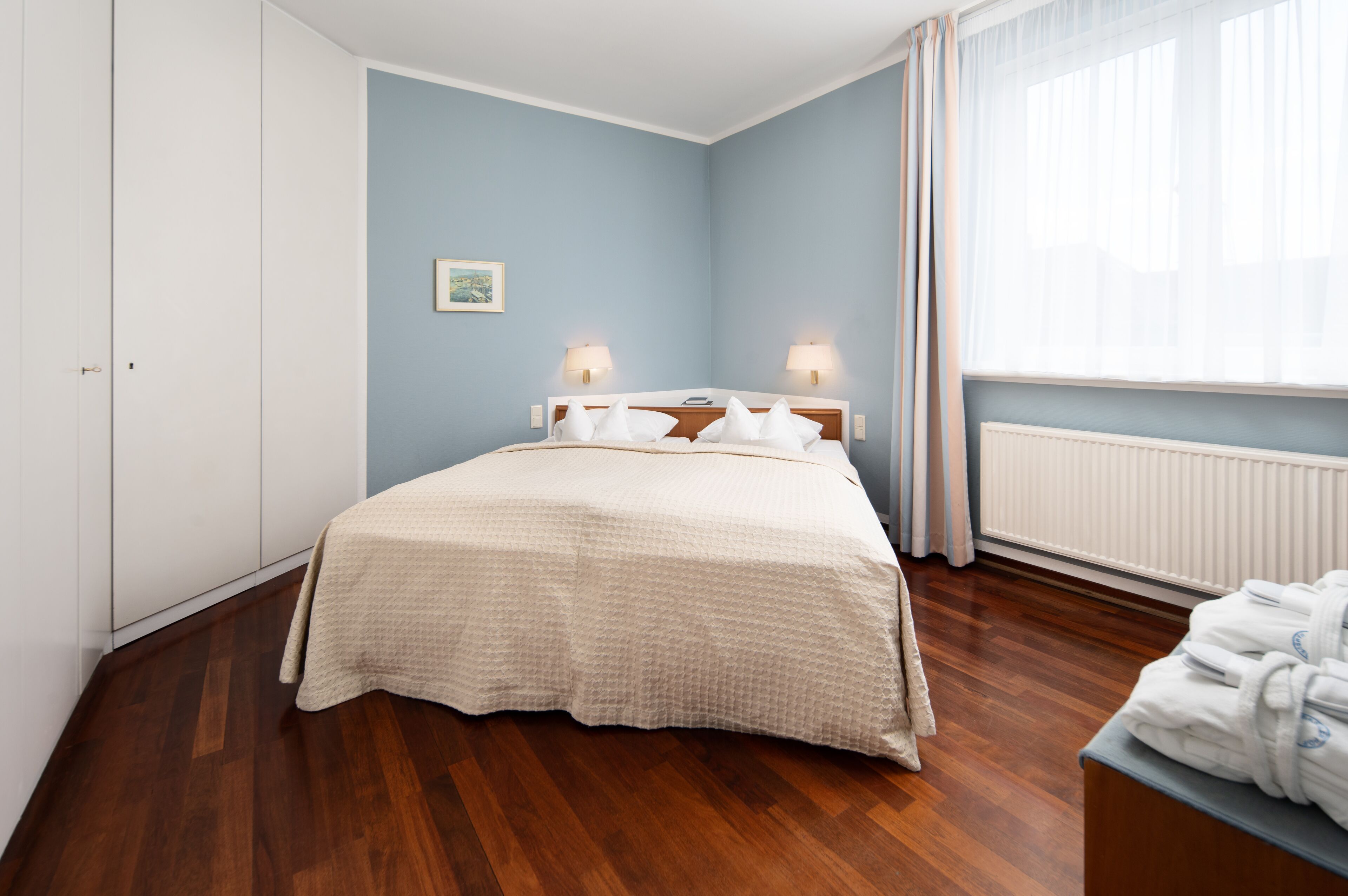 A bedroom with a bed, two bedside lamps, a radiator, and a window with curtains.