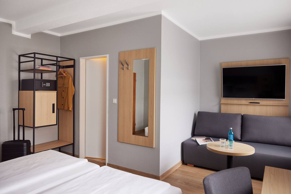 A hotel room with a bed, a sofa, a TV, a small table, and a wardrobe with a mirror.