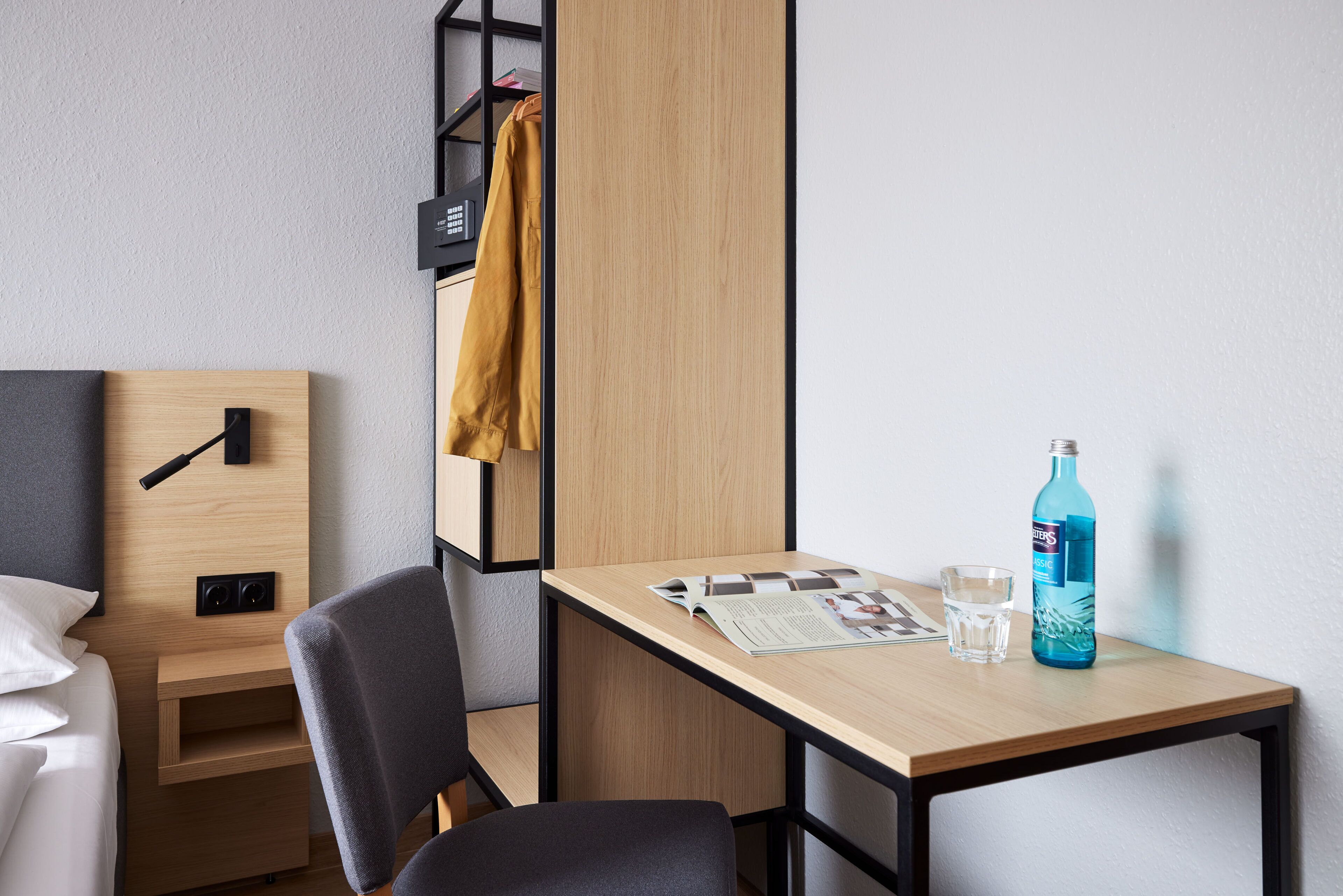 A hotel room with a bed, a desk with a magazine, a bottle of water, and a chair.