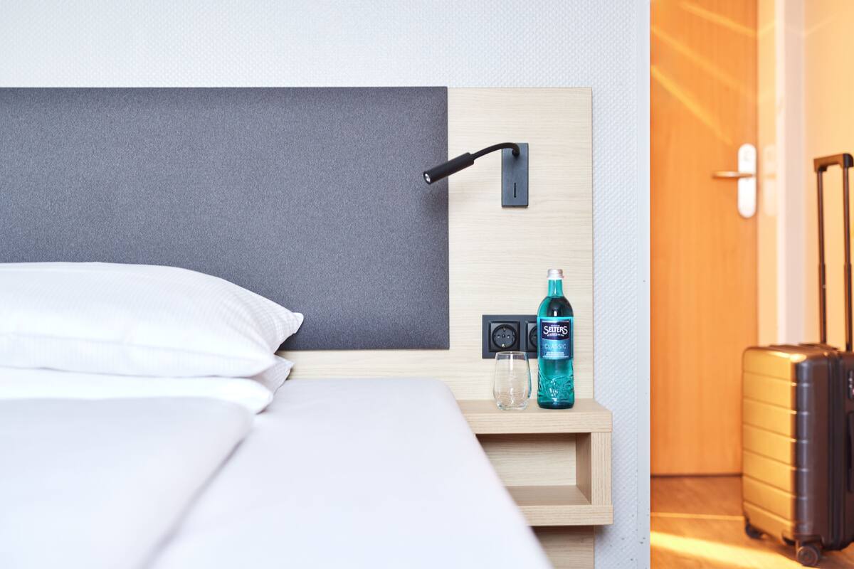 A hotel room with a bed, a bedside table with a bottle of hand sanitizer, a lamp, and a suitcase.