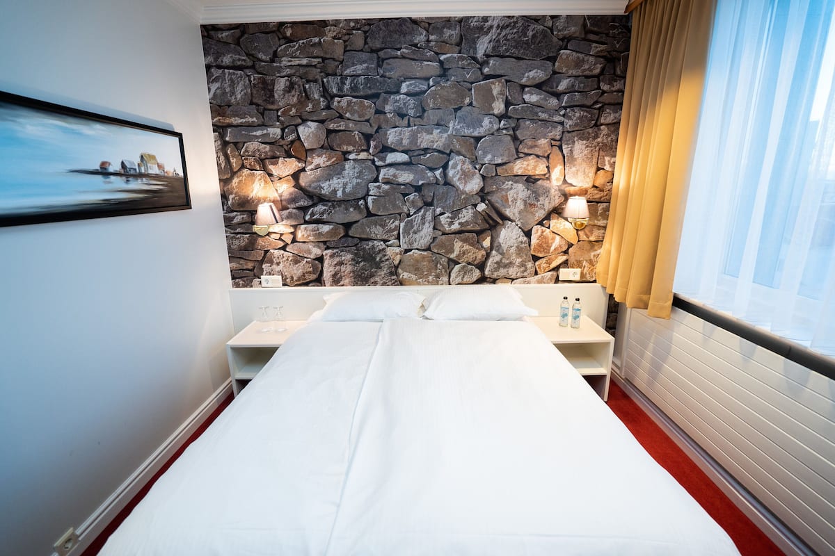 Ein Hotelzimmer mit Steinwand, einem Bett mit weißen Leinentüchern, einem Nachttisch mit zwei Flaschen und einem Bild an der Wand.