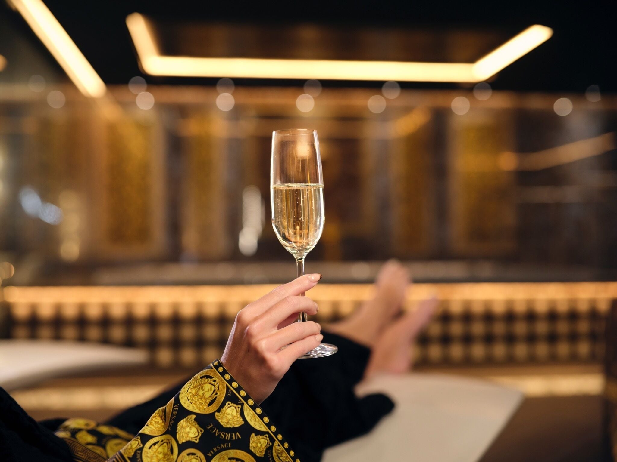 Uma pessoa relaxada, reclinada com uma taça de champanhe, segurando uma manga com padrão dourado, em um ambiente com iluminação suave e decoração moderna.