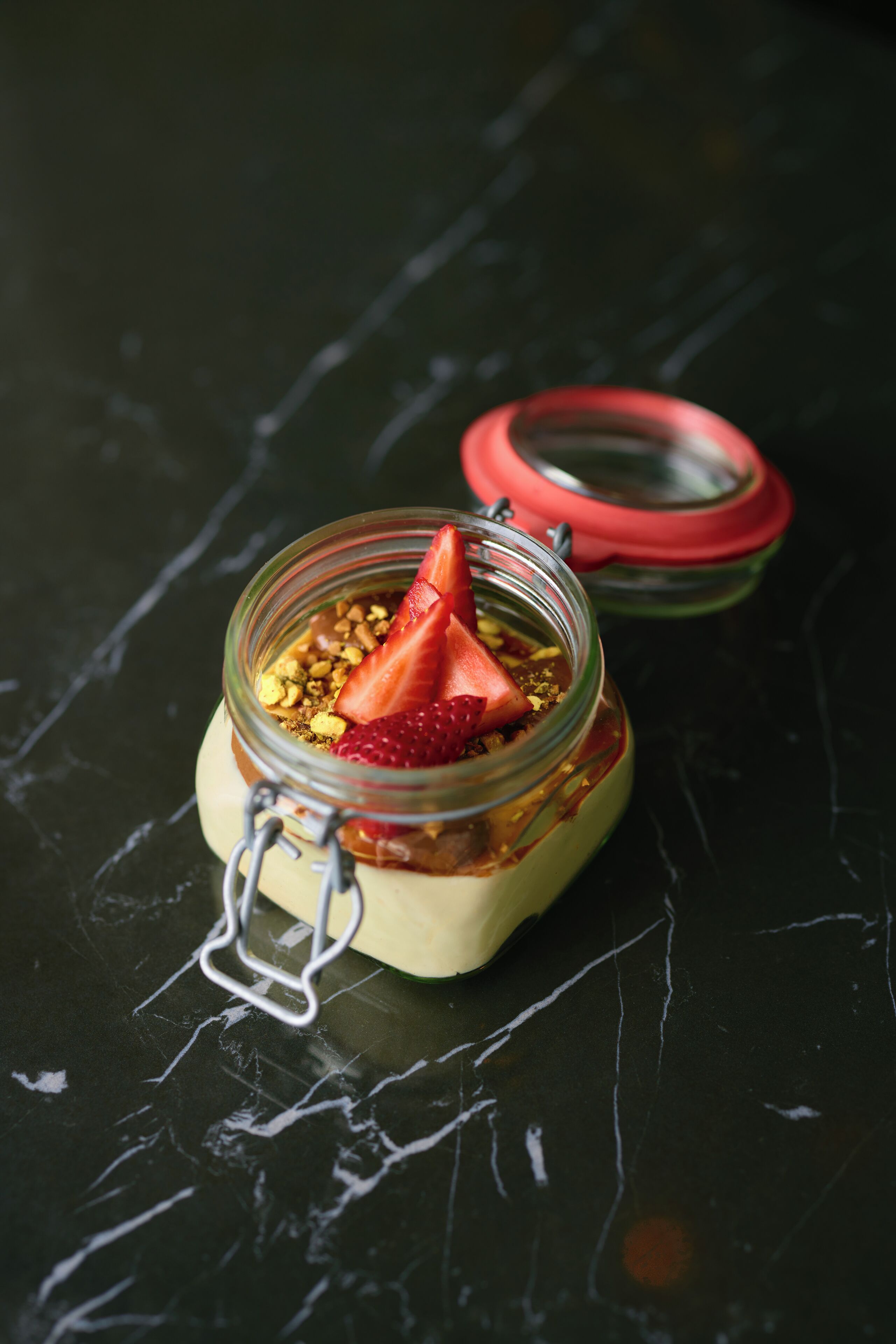 Ein Glasgefäß mit Schraubverschluss, gefüllt mit einer geschichteten Nachspeise mit Erdbeeren und Granola auf einer dunklen Oberfläche.