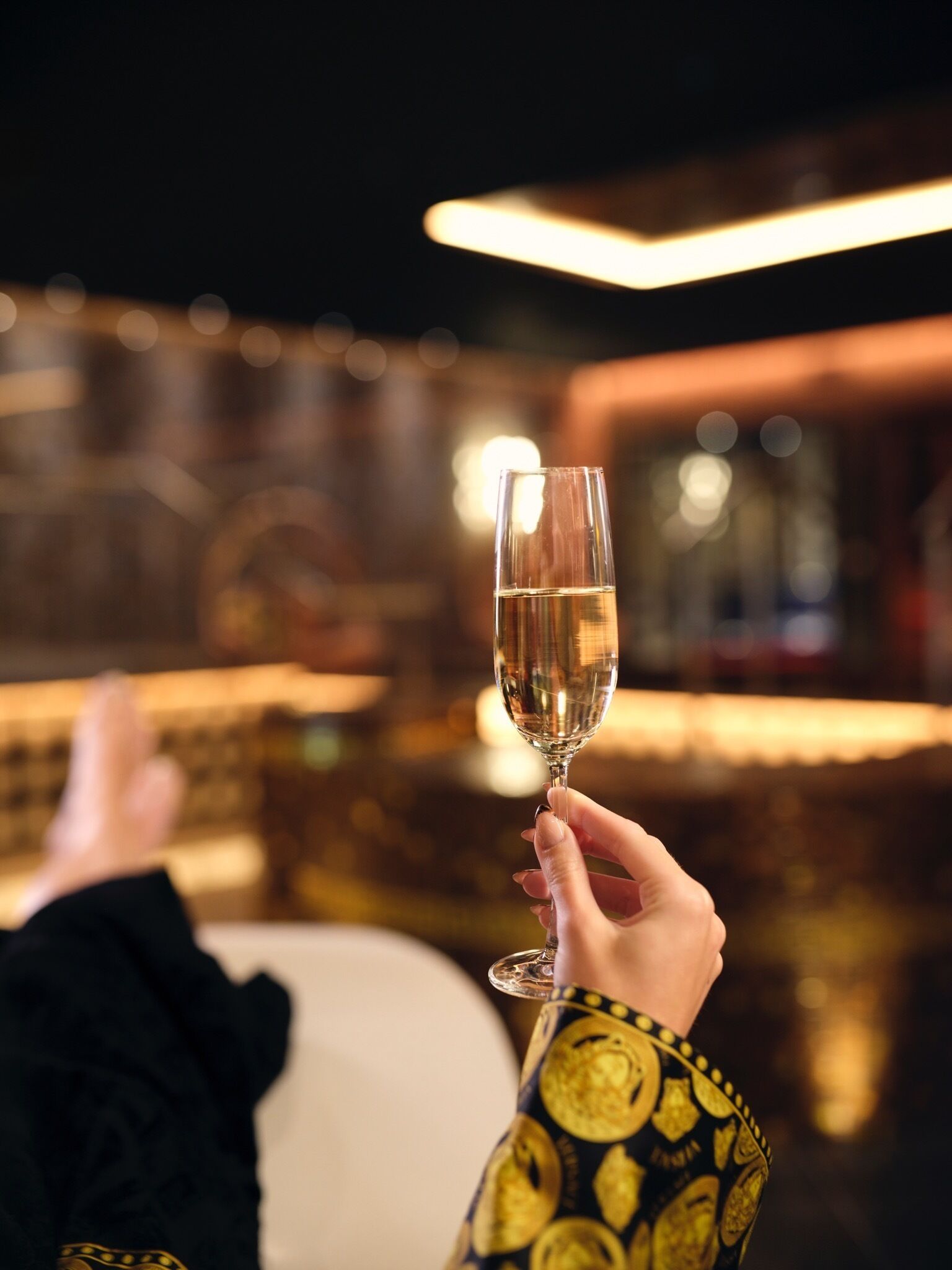 A person holding a champagne glass, reclining on a white sofa in a dimly lit room with a bar in the background.