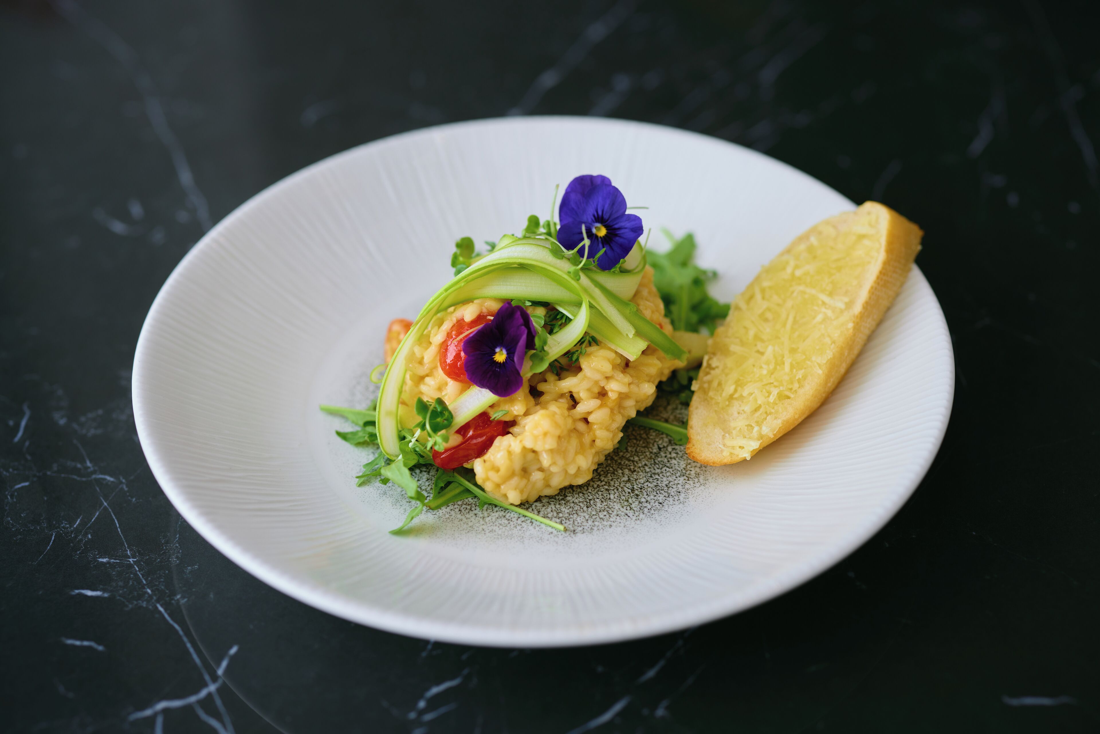 A white plate with a dish of risotto, garnished with green onions, purple flowers, and red tomatoes, accompanied by a slice of bread on a dark surface.