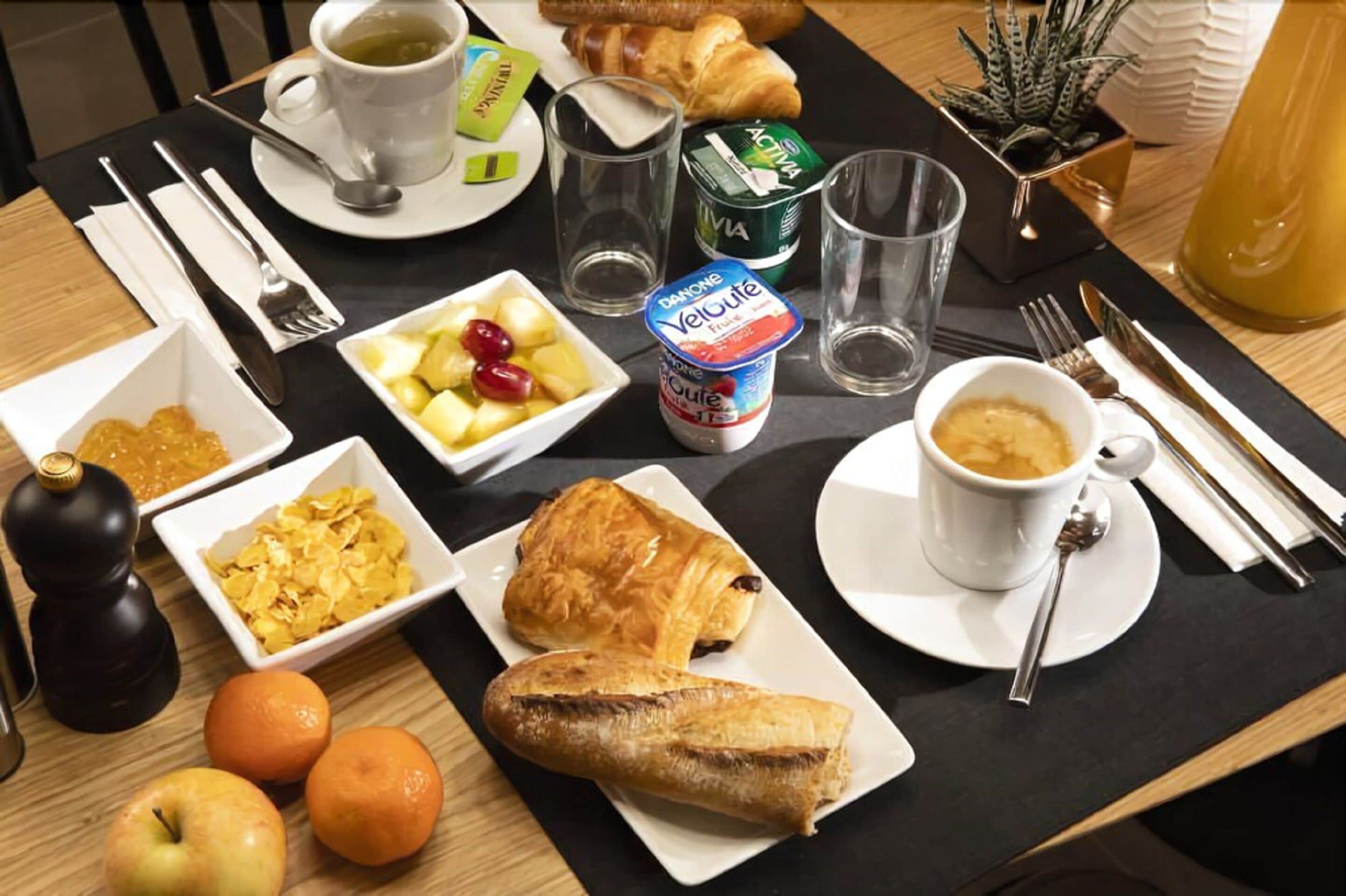 Una mesa de desayuno con cruasanes, frutas, yogur, café y una botella de jugo.