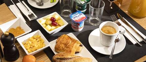 Una mesa de desayuno con cruasanes, frutas, yogur, café y una botella de jugo.