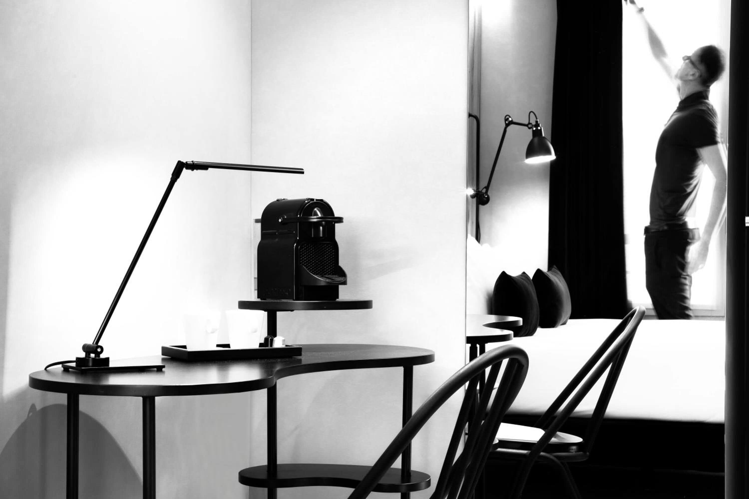 Una habitación moderna con una mesa redonda, una lámpara y una silla. Hay una ventana con cortinas, y se ve a una persona a través de ella.