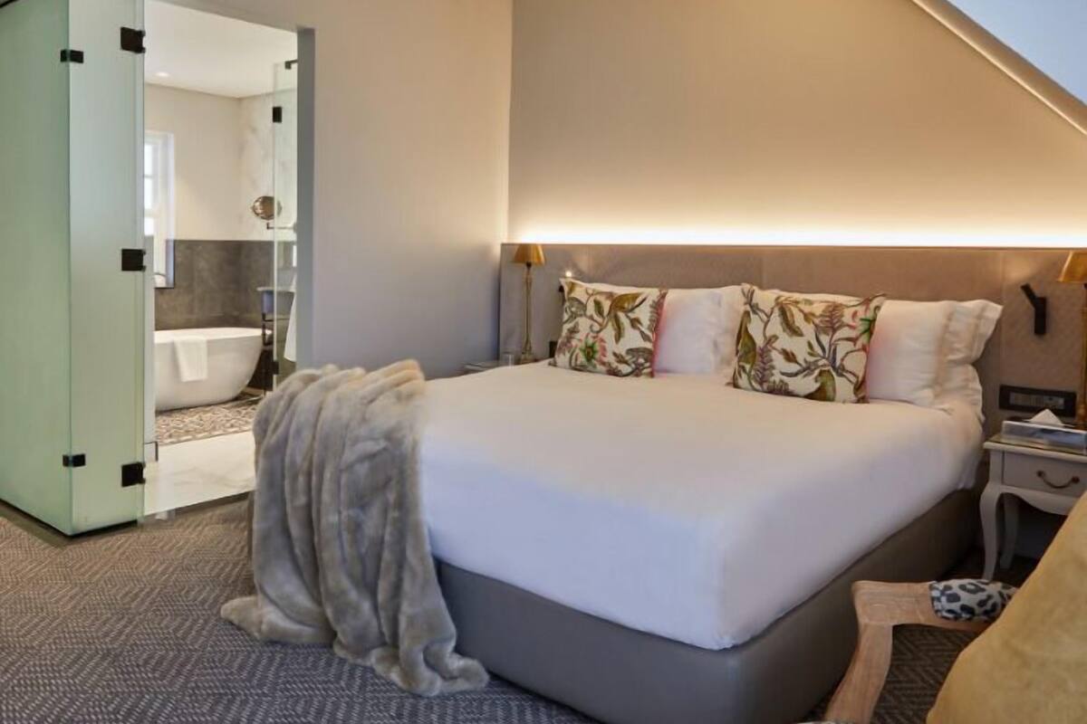 A modern hotel room with a large bed, a bedside table, a nightstand, and a bathroom visible through a glass door.