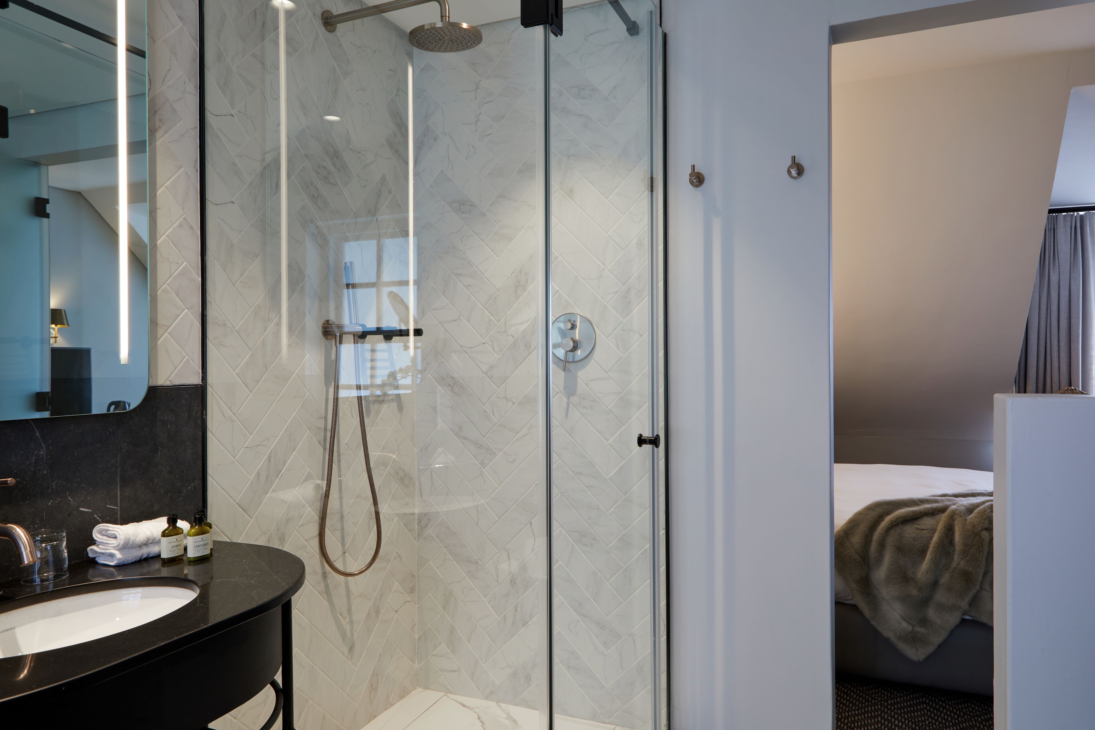 A modern bathroom with a glass-enclosed shower, a black vanity with a mirror, and a bed with a gray blanket in the adjacent room.