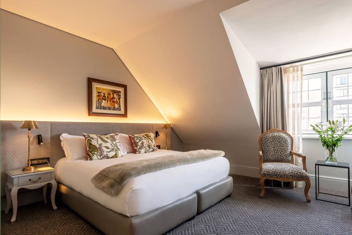 A hotel room with a large bed, a chair, a small table, a painting, and a window with curtains.