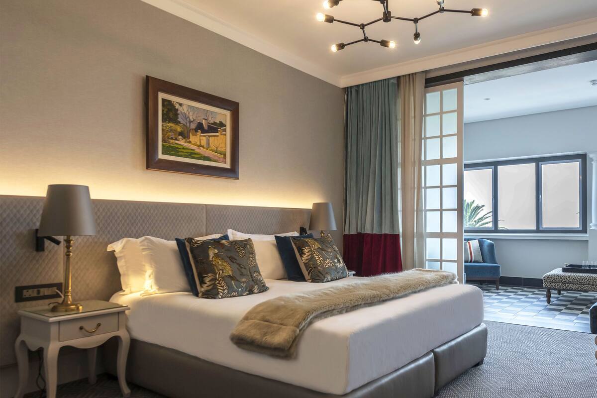A modern hotel room with a large bed, a nightstand, a lamp, a painting on the wall, and a view of the outdoors through a window.