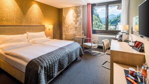 A hotel room with a large bed, a desk with a computer, a chair, and a view of mountains through the window.