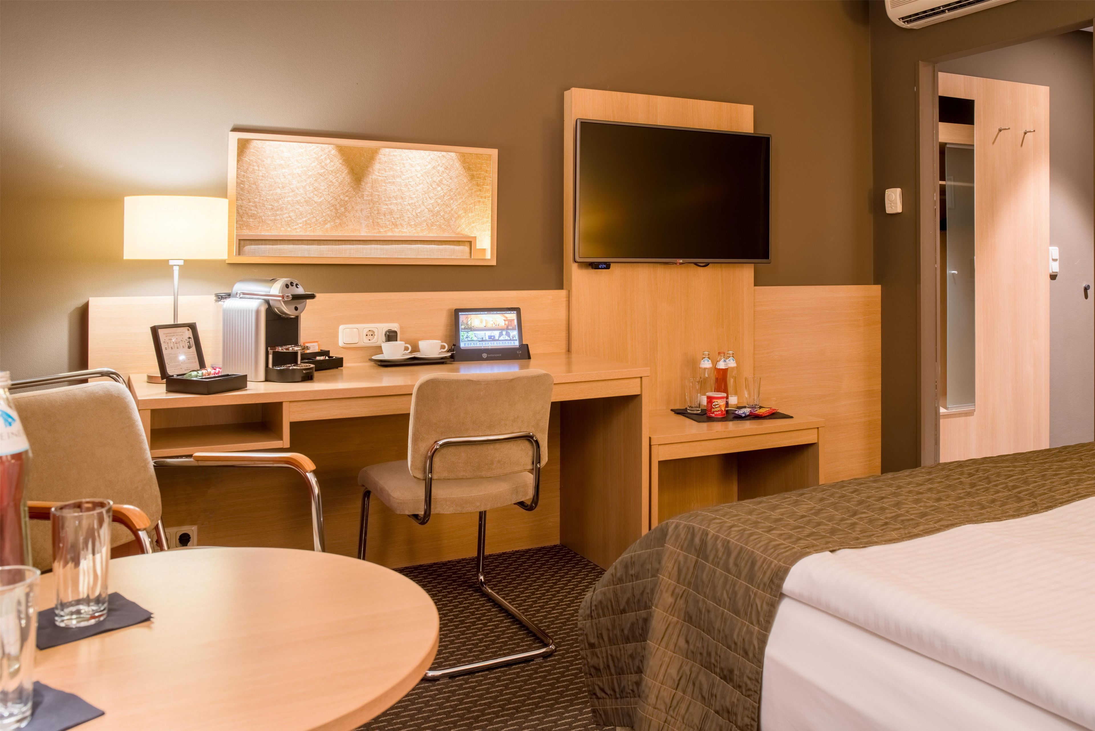 A hotel room with a desk, chair, bed, TV, and a minibar.