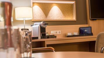 A hotel room with a desk, a coffee machine, a laptop, a TV, and a mirror.