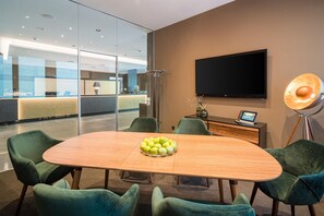 Une salle de conférence moderne, dotée d’une table en bois, de chaises recouvertes d’un tissu vert, d’un téléviseur à écran plat et d’un bureau avec une lampe et une tablette.