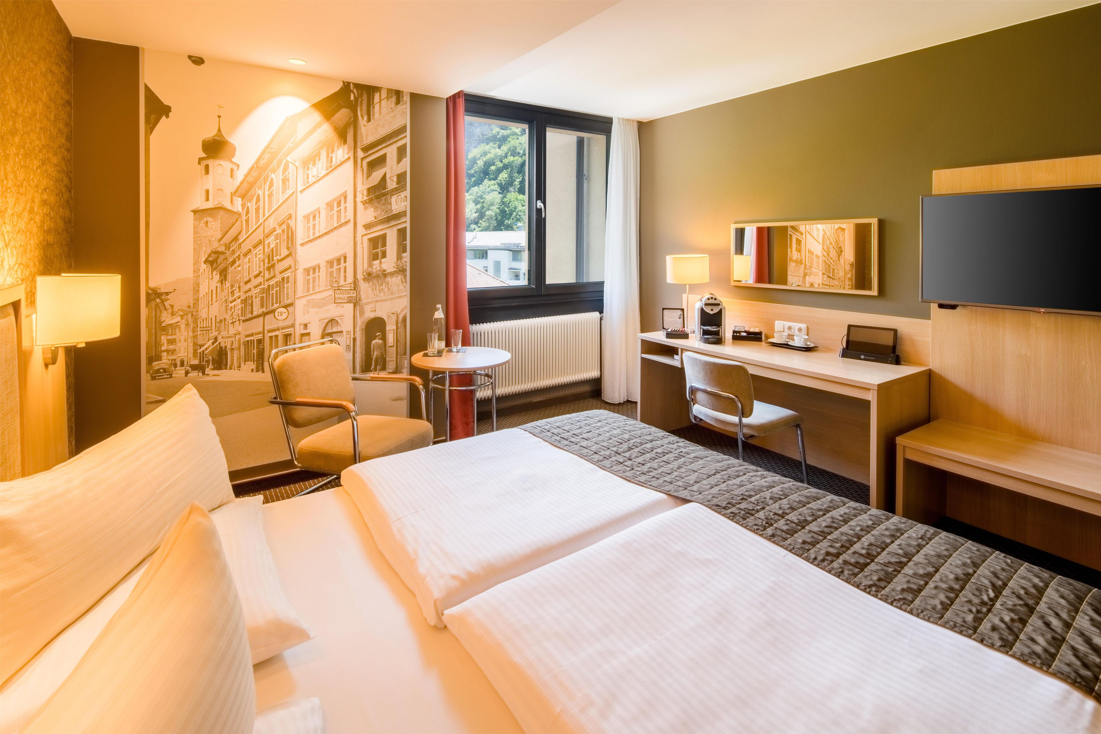 A hotel room with a bed, a desk, a chair, a TV, and a view of buildings through the window.