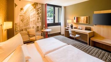 A hotel room with a bed, a desk, a chair, a TV, and a view of buildings through the window.