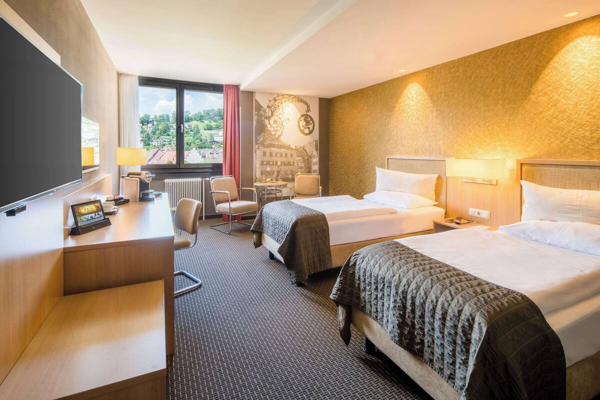 Een hotelkamer met twee bedden, een bureau, een stoel, een televisie en uitzicht op de omgeving.
