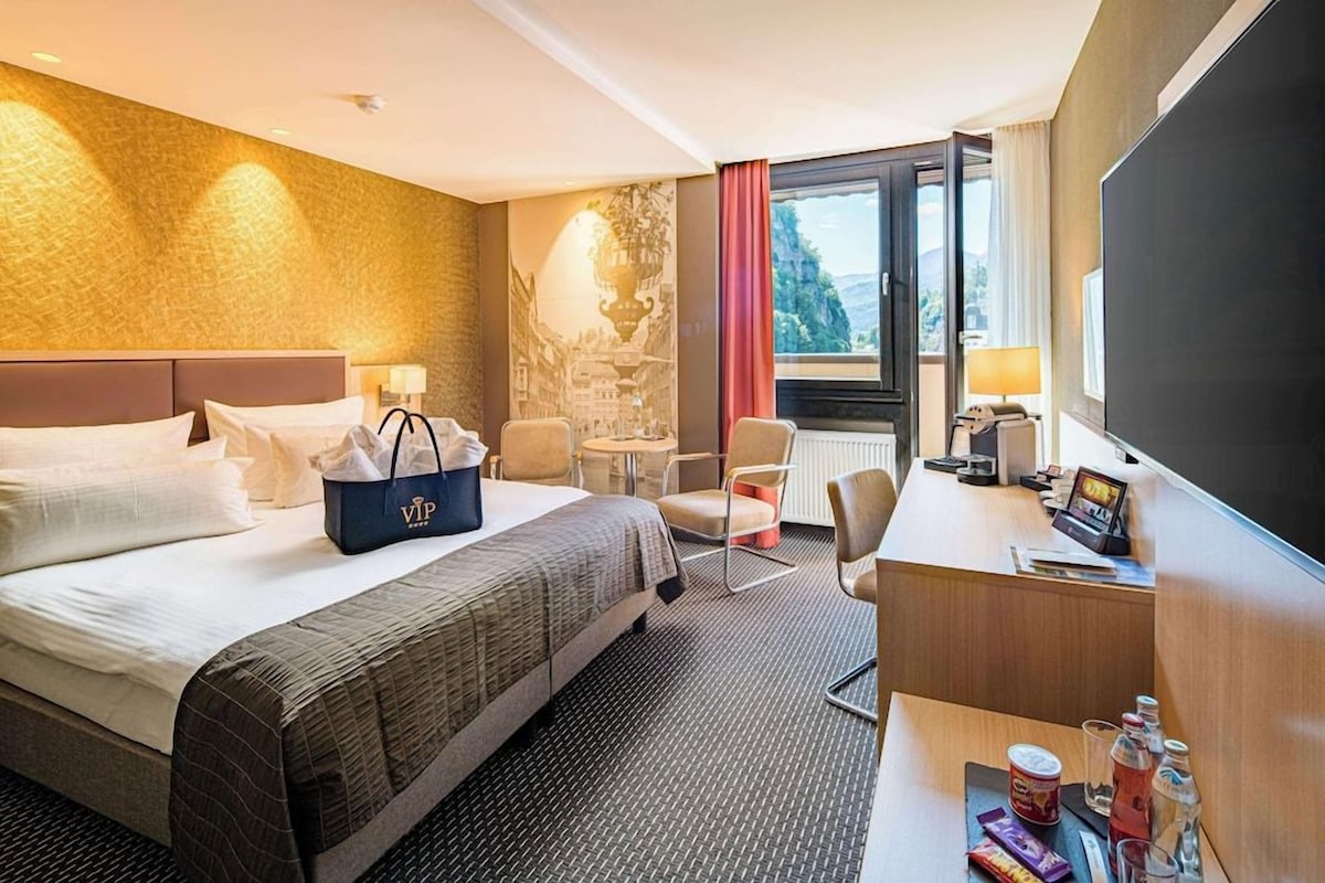 Habitación de hotel con cama, escritorio, silla, bolso y vista al exterior.