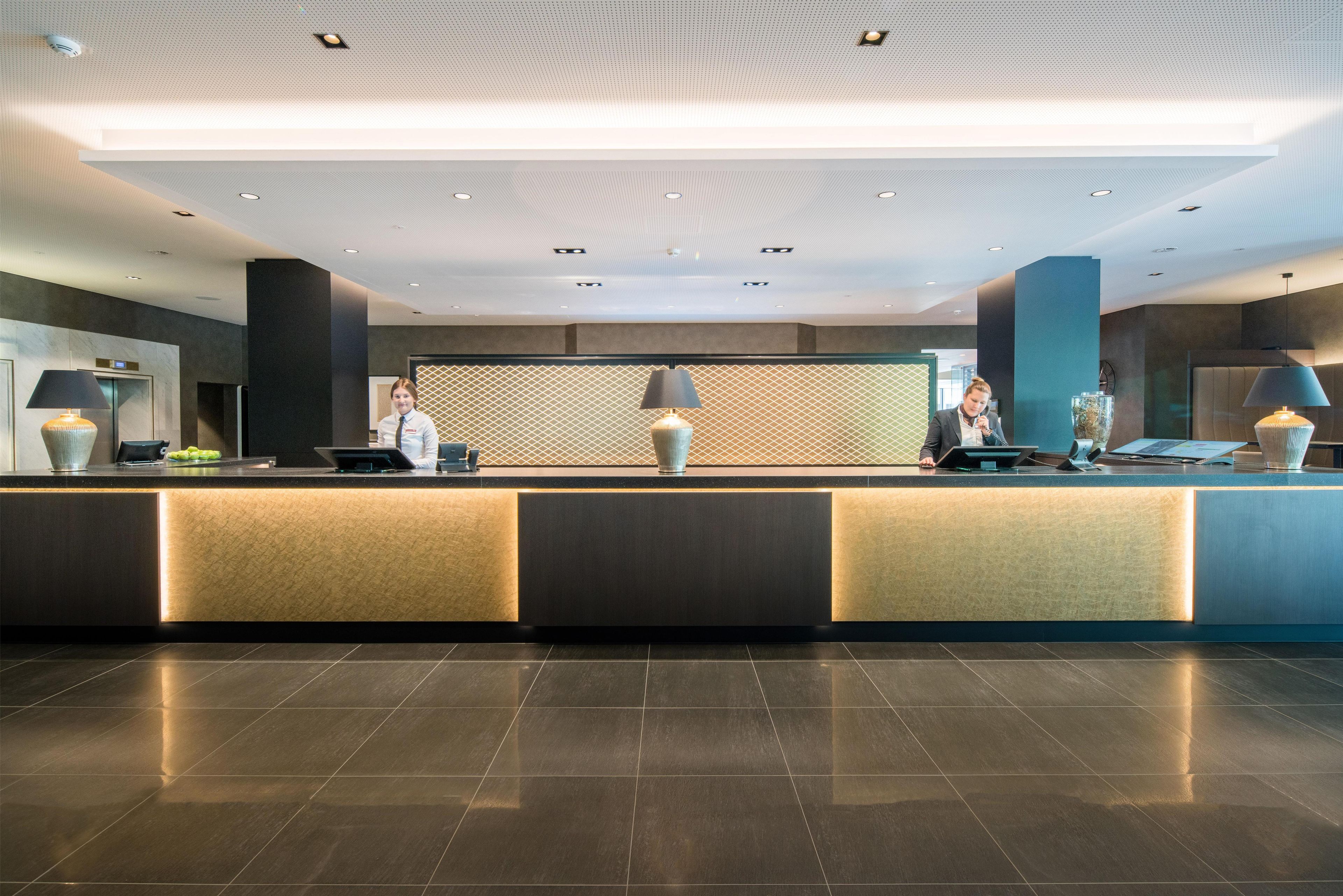 A hotel reception area with a modern design, featuring a reception desk, two receptionists, a patterned wall, and a ceiling with recessed lighting.