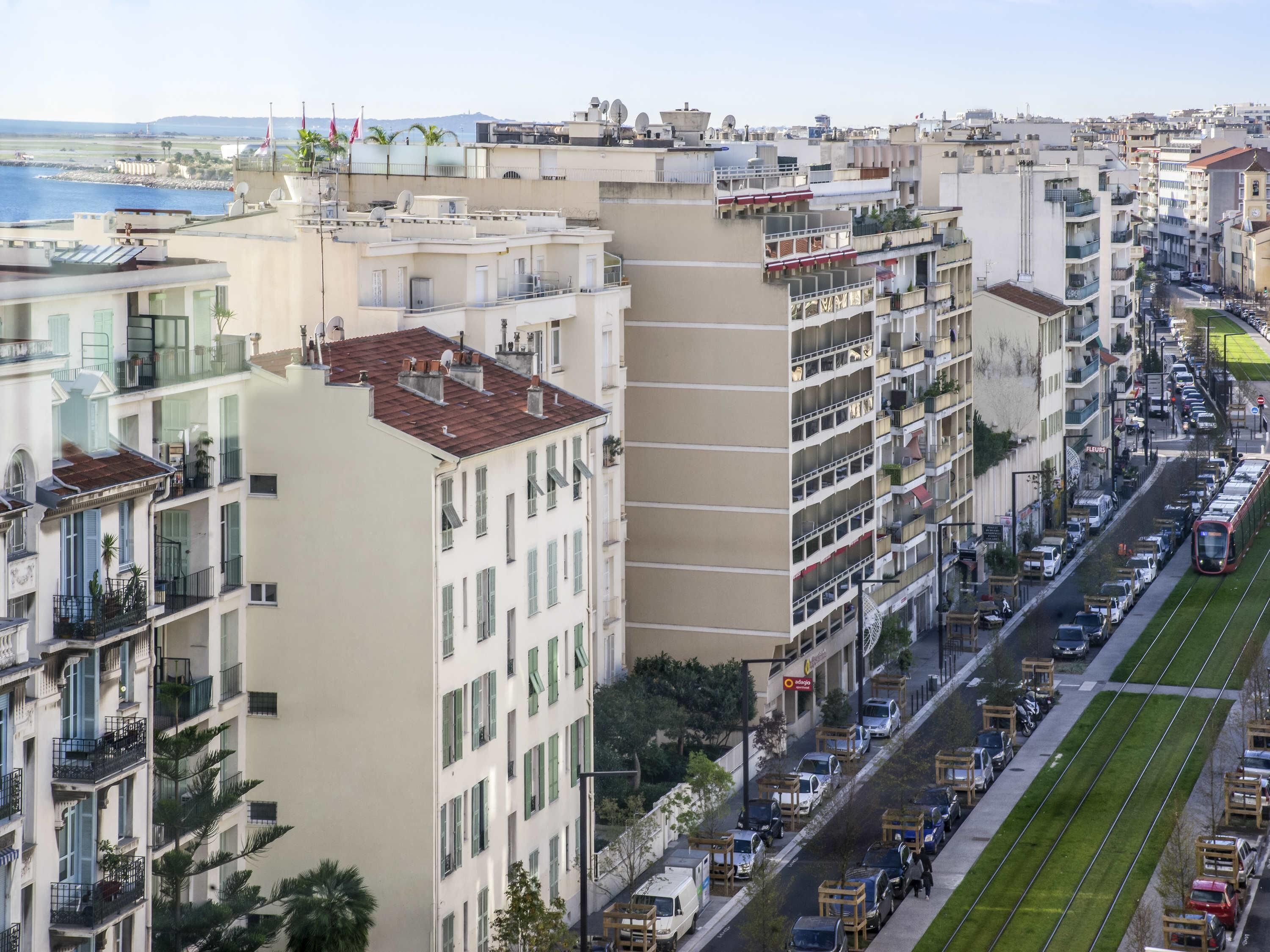 Un edificio de varios pisos con balcones, una calle con coches aparcados y una línea de tranvía.
