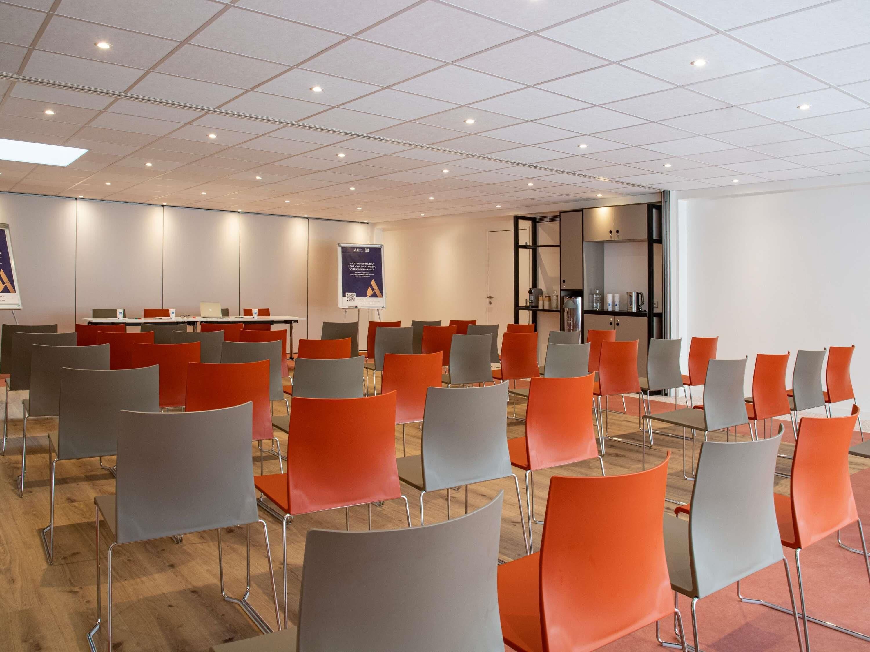A conference room with rows of chairs, a presentation screen, and a kitchenette area.