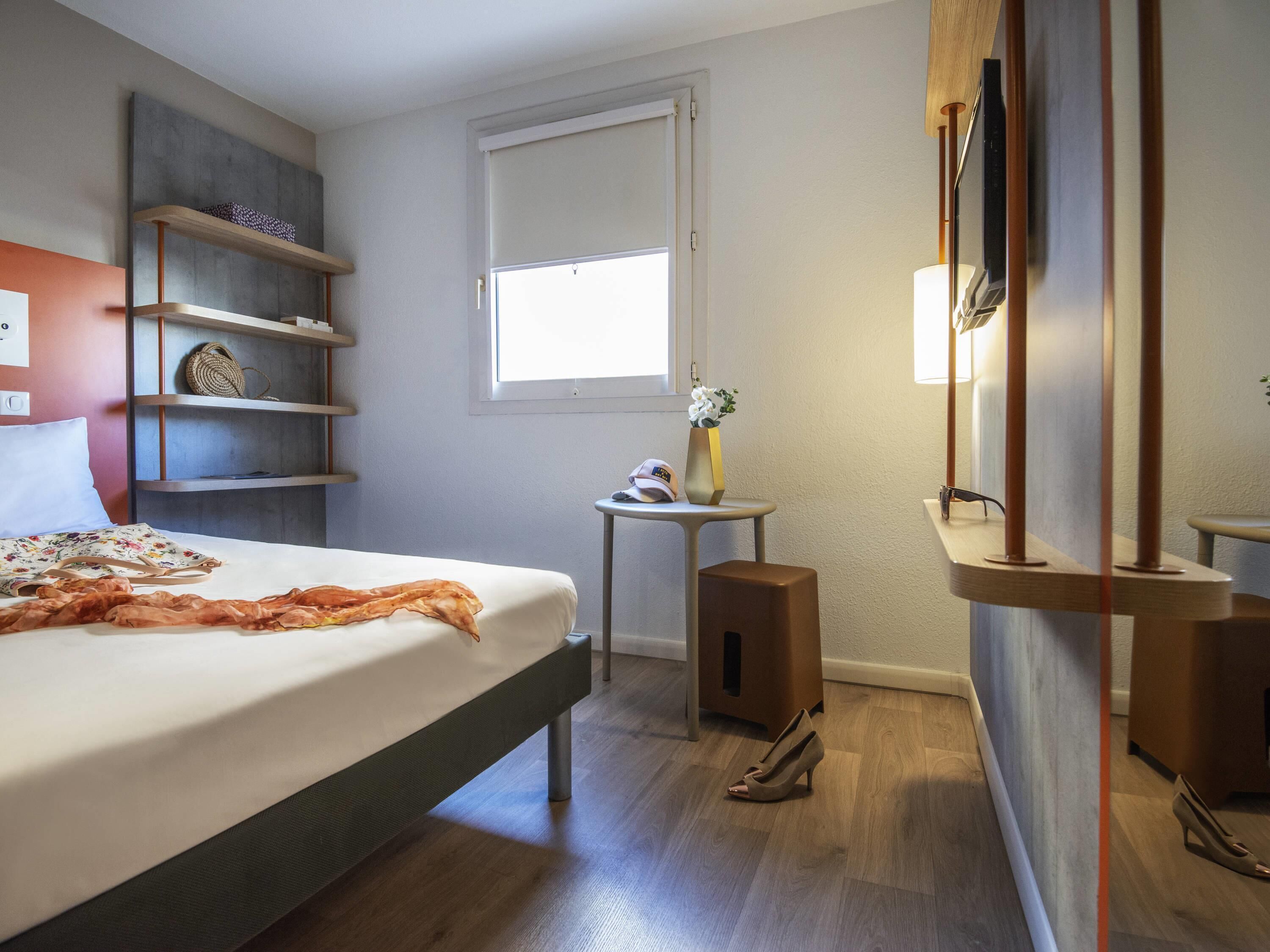 Una habitación de hotel con una cama, una mesita con un florero y un gorro, un espejo, un altavoz y una repisa con una cesta tejida.
