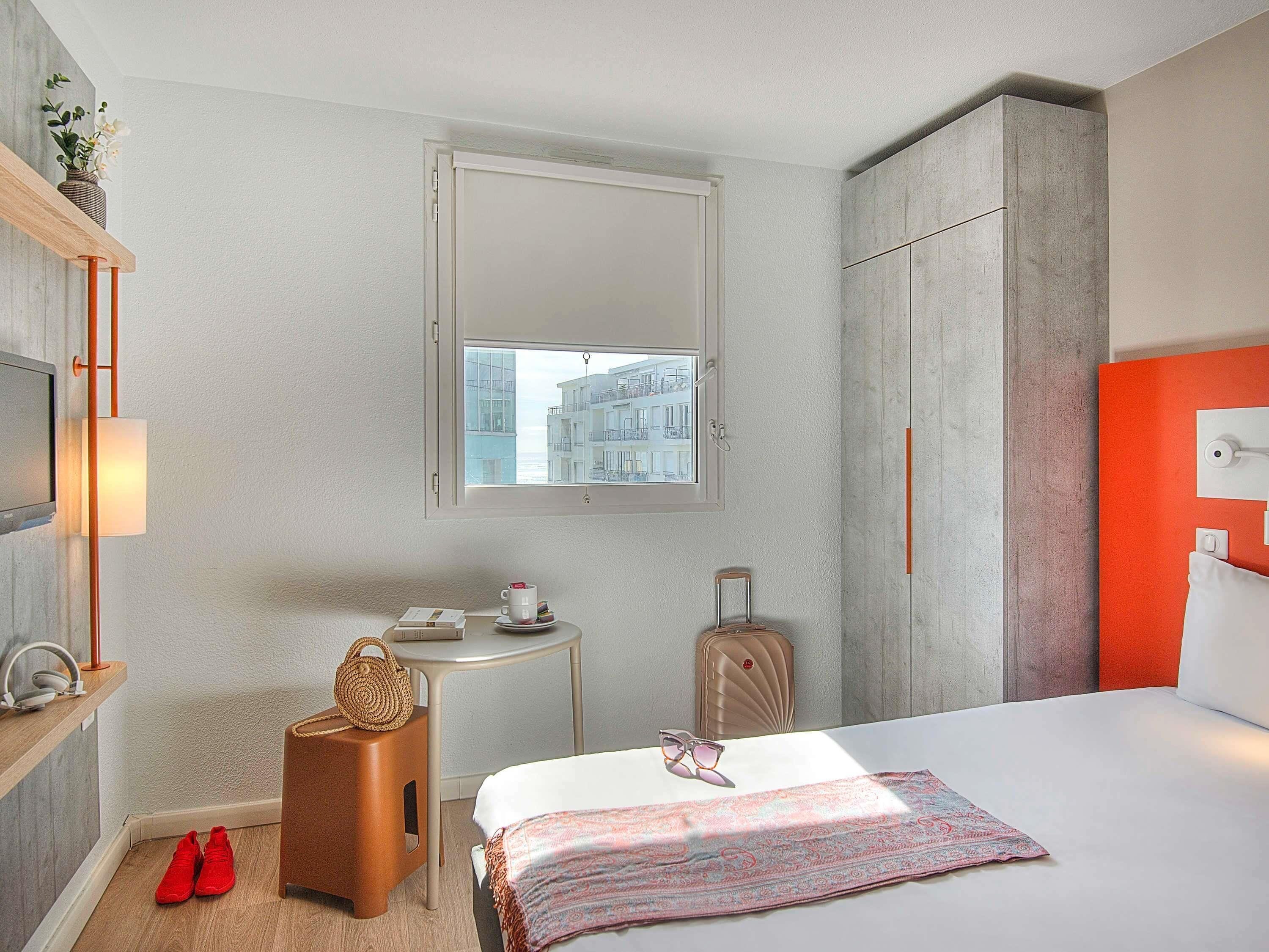 Habitación de hotel con cama, una mesita, una silla, una maleta, televisión y ventana con vistas a edificios.