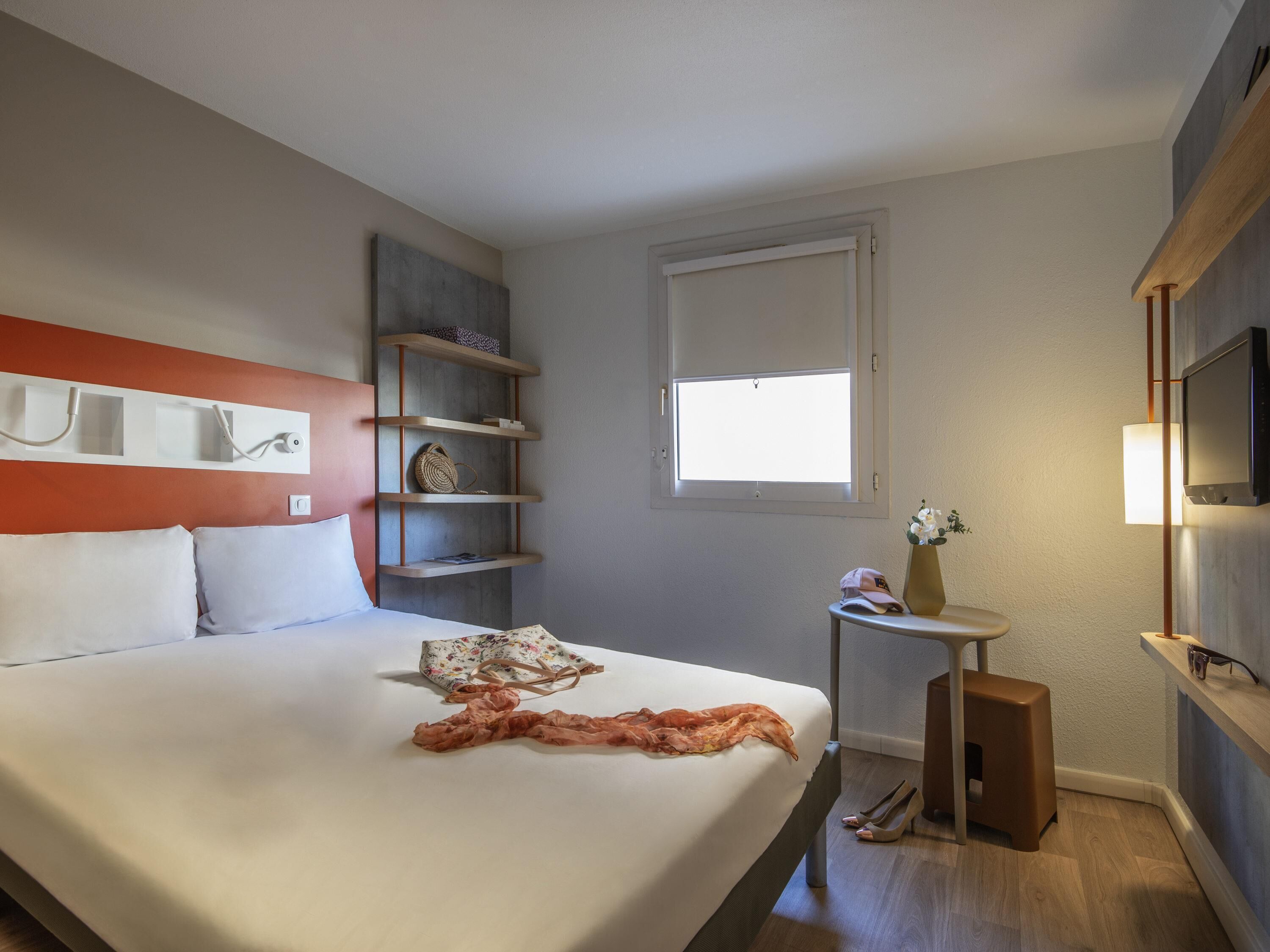Ein Hotelzimmer mit Bett, Fernseher, Fenster, kleinem Tisch und einer Ablage mit Gegenständen.
