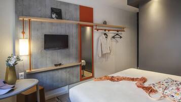 Ein modernes Hotelzimmer mit Bett, einem an der Wand befestigten Fernseher, einem Spiegel, einem kleinen Tisch mit Stuhl und einem Kleiderständer.