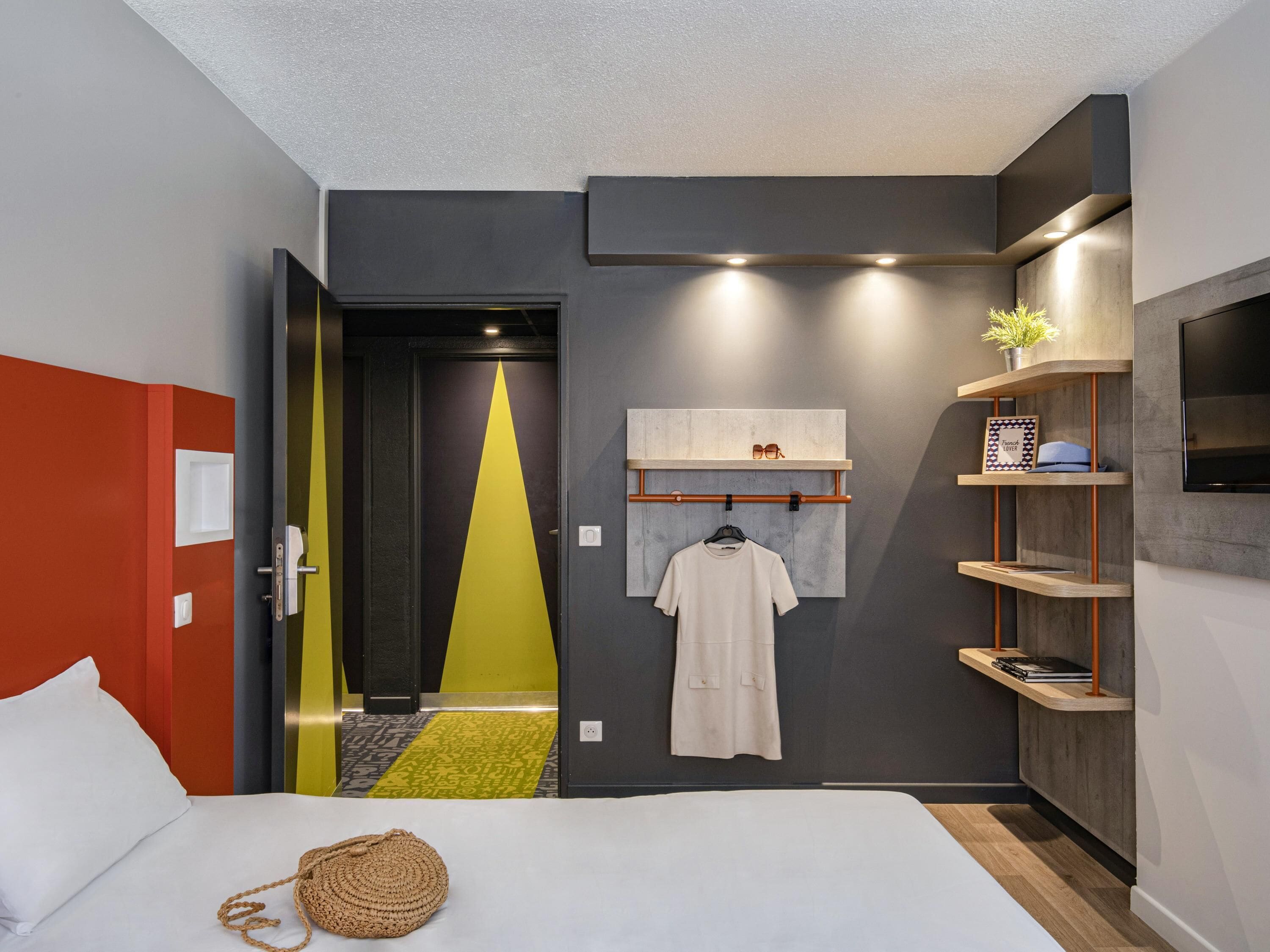 Une chambre d’hôtel moderne équipée d’un lit, d’une robe suspendue, d’une étagère avec des livres et d’un téléviseur.