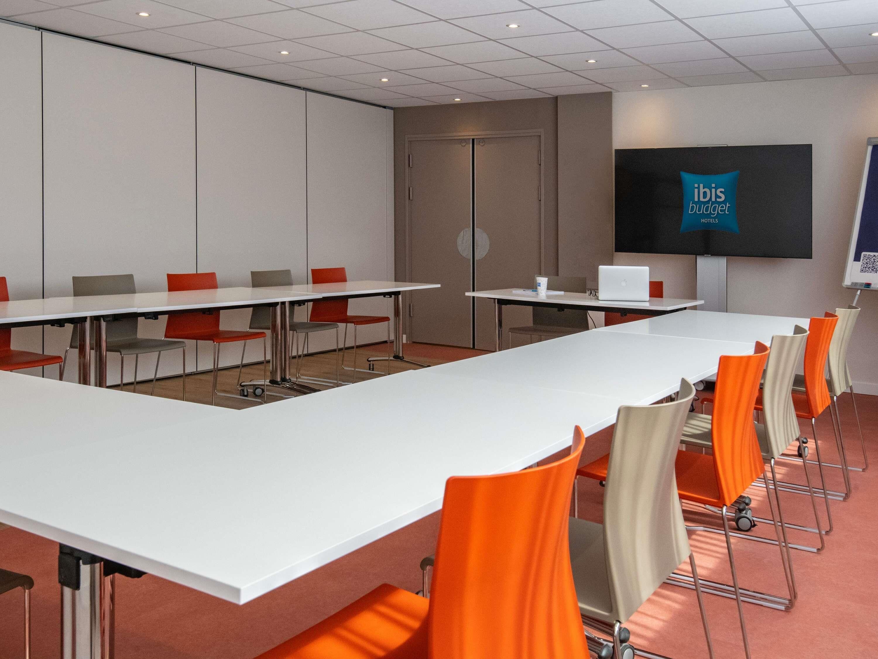 Un salón de conferencias con una larga mesa, sillas, una pantalla de proyector y una computadora portátil.