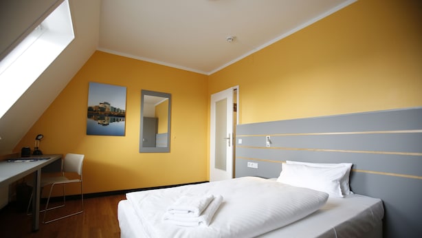 A hotel room with a bed, a desk, a chair, a mirror, and a painting on the wall. - Dietrich-Bonhoeffer-Hotel (Berlin)