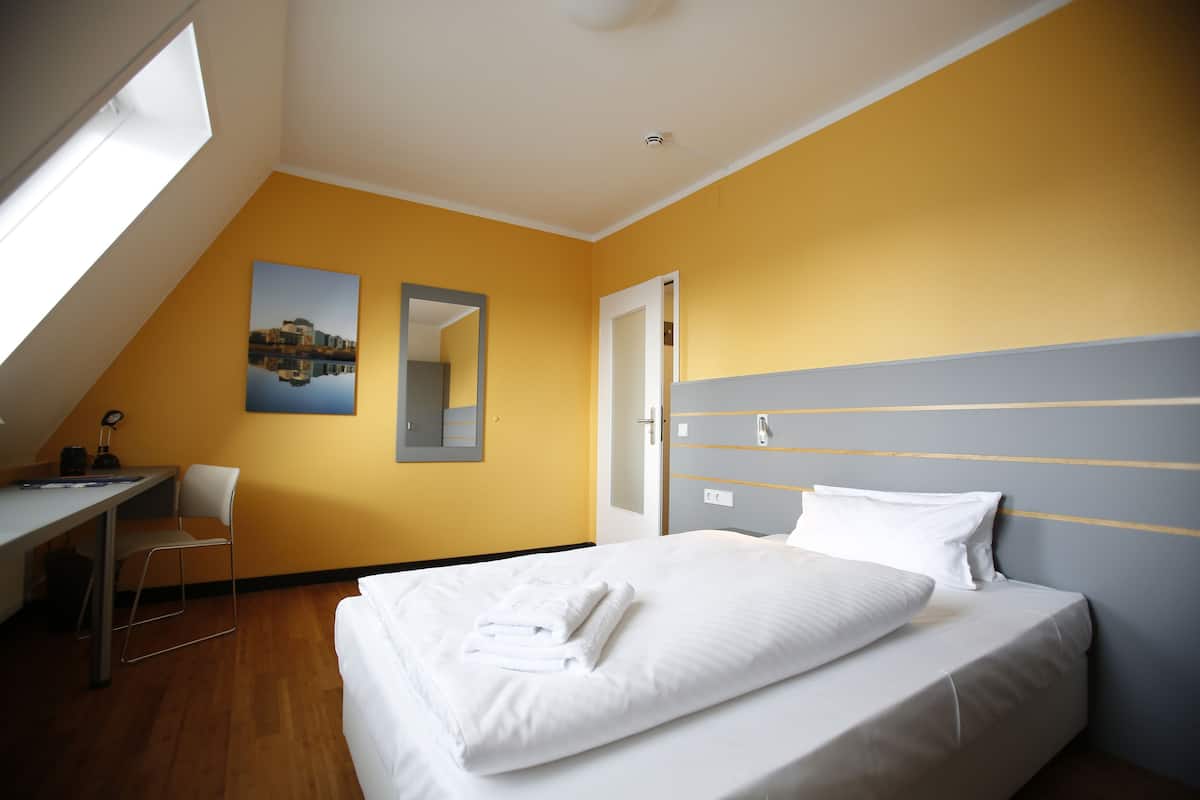 A hotel room with a bed, a desk, a chair, a mirror, and a painting on the wall.