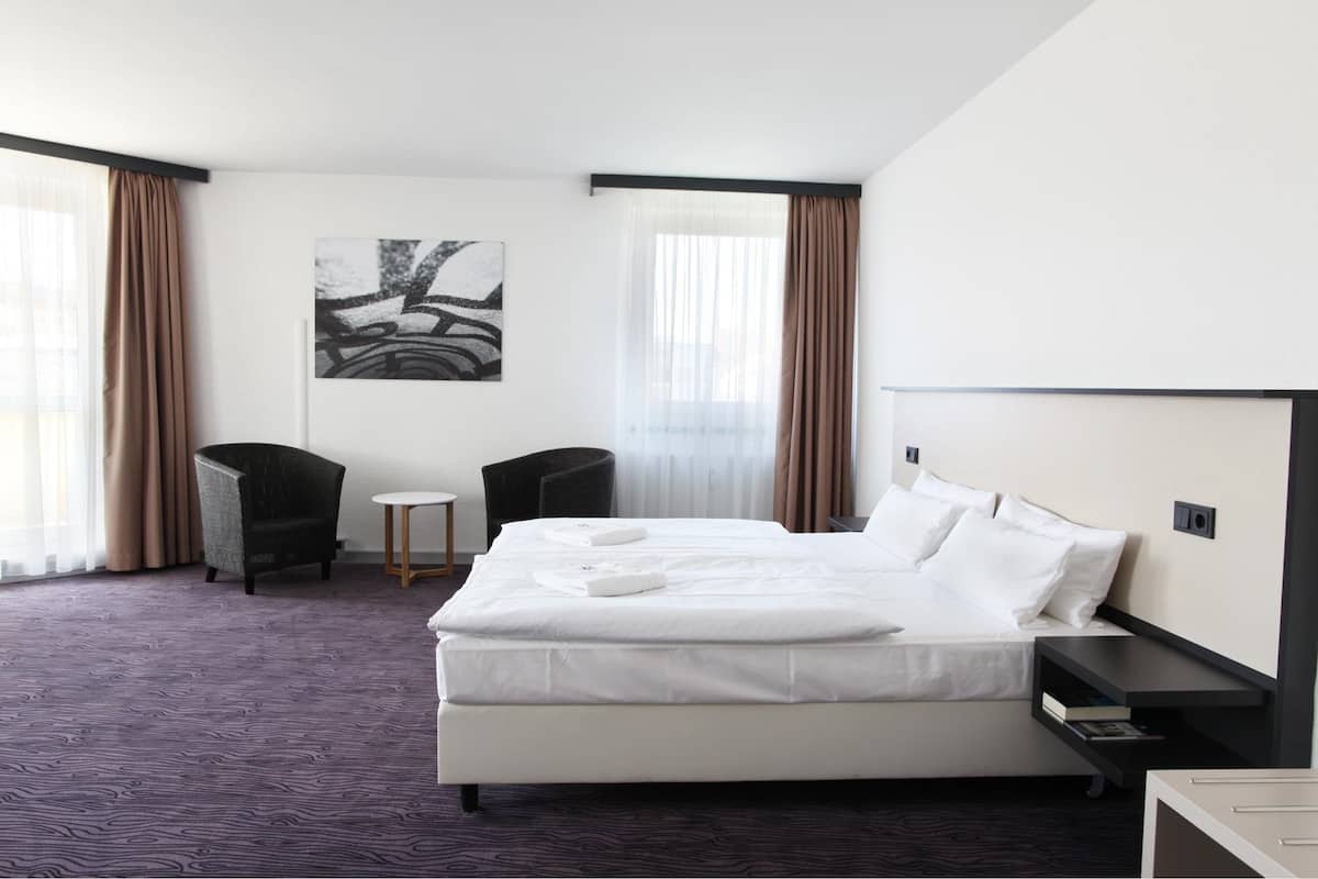 A hotel room with a large bed, a small table, two chairs, a painting on the wall, and a window with curtains.