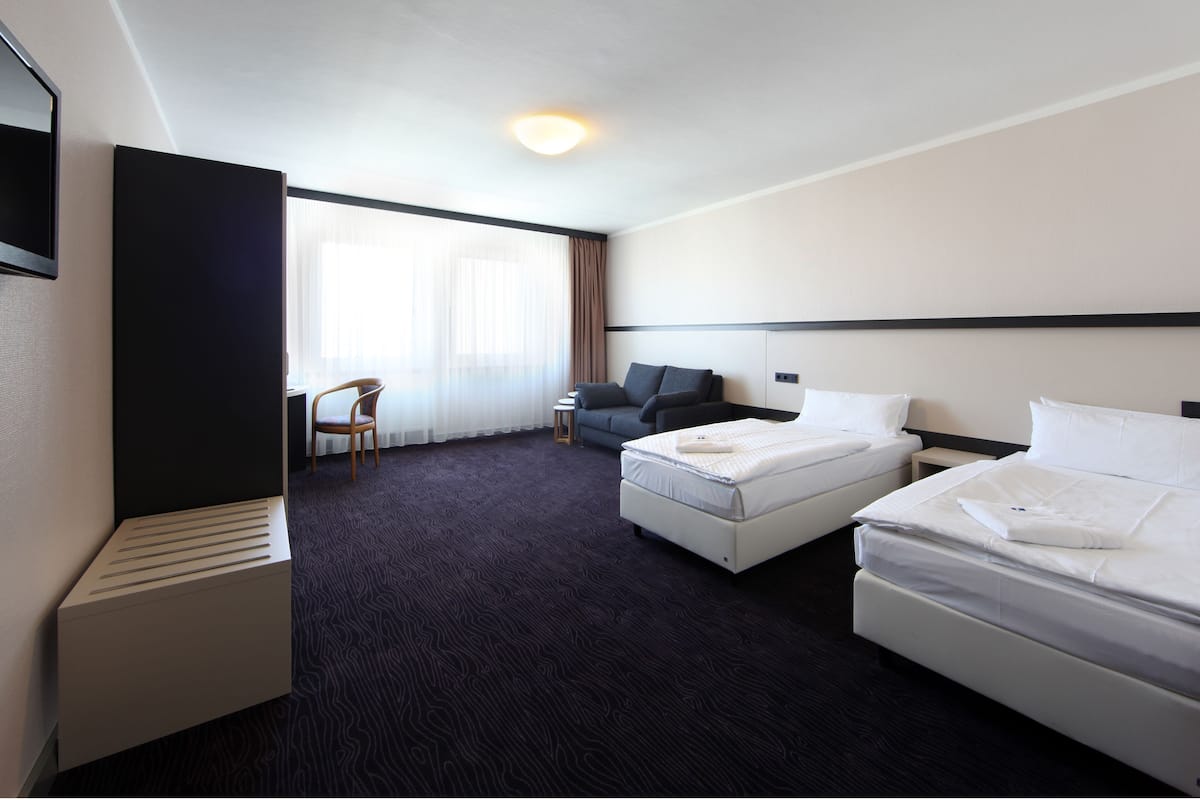 A hotel room with two beds, a desk, a chair, and a large window with sheer curtains.