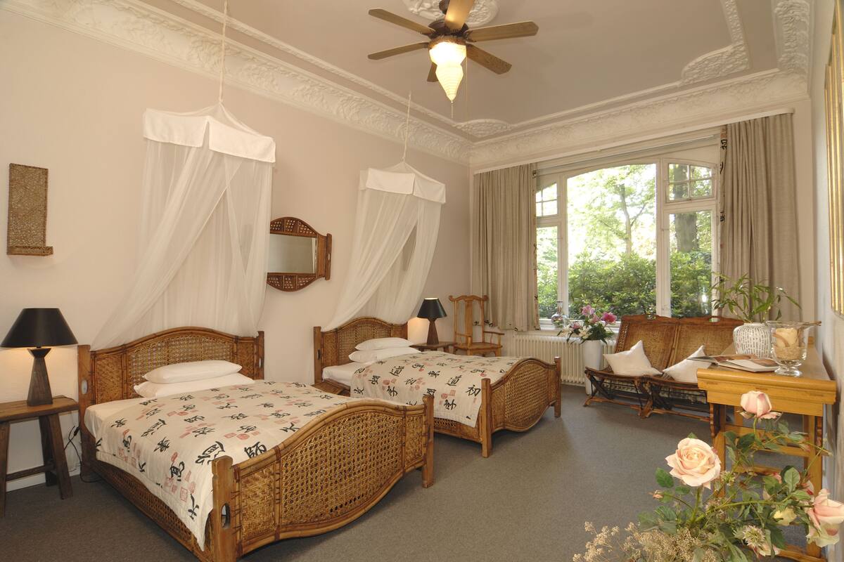Ein Hotelzimmer mit zwei Holzbett, einem Deckenventilator, einem großen Fenster mit Vorhängen, einem Nachttisch mit Lampe und einer Vase mit Blumen.