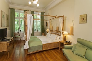 A four-poster bed with a canopy, a green sofa, a wooden desk with a lamp, and a television in a room with wooden flooring and green curtains. - Hotel Miramar (Hamburg)