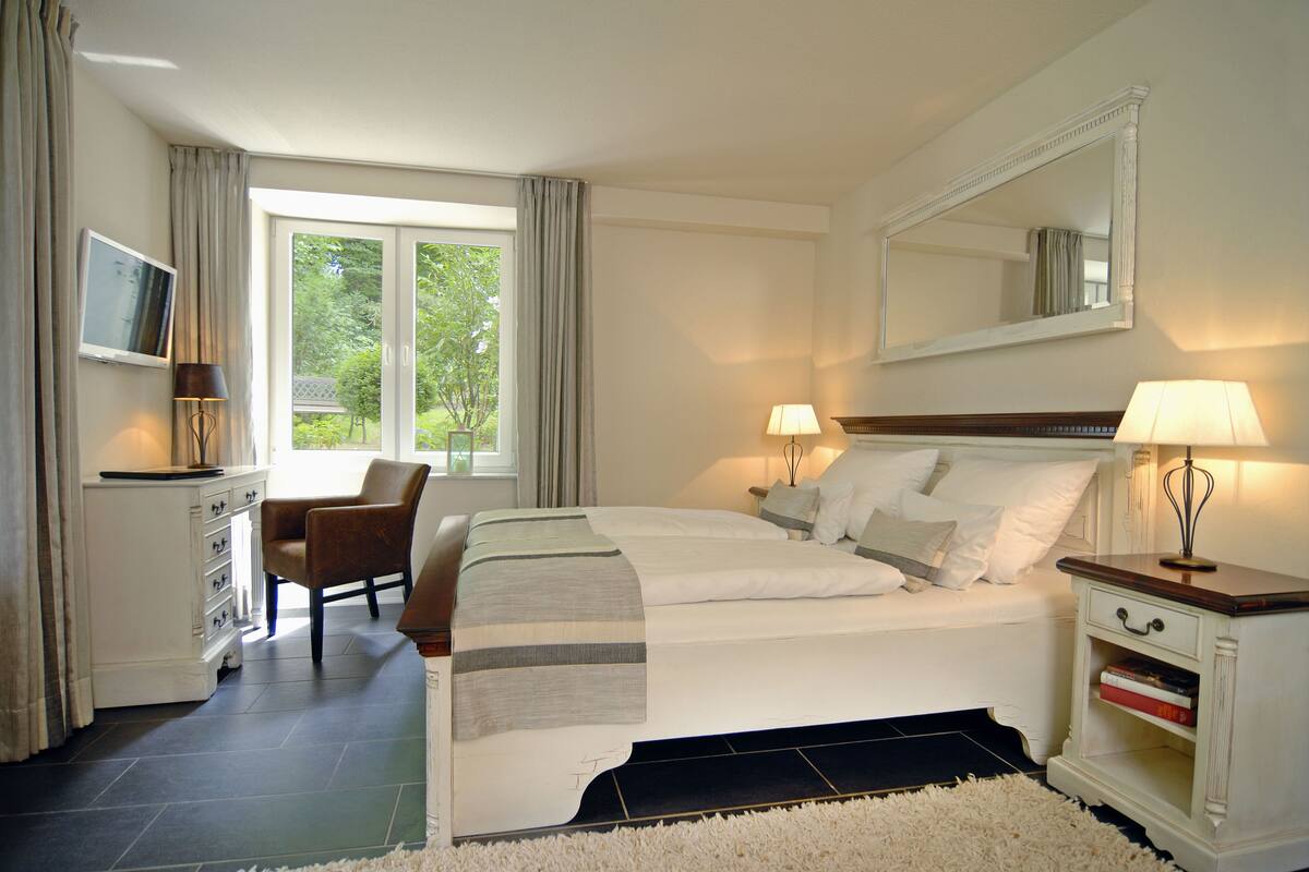 Ein Hotelzimmer mit Bett, Sessel, Fernseher, Schreibtisch und einem Fenster mit Vorhängen.