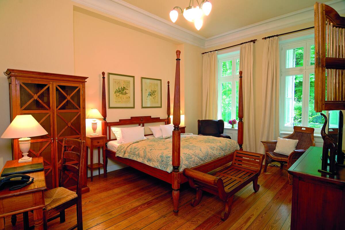 Ein traditionell eingerichtetes Schlafzimmer mit Himmelbett, Holzmöbeln und einem großen Fenster mit Vorhängen.