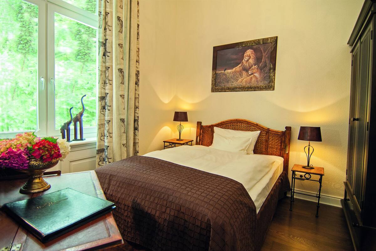 Ein ordentlich bezogenes Bett mit einer braunen Steude, zwei Nachttischlampen, ein gerahmtes Bild an der Wand, ein Fenster mit Vorhängen und ein Schreibtisch mit einer Blumenvase und einem Buch.