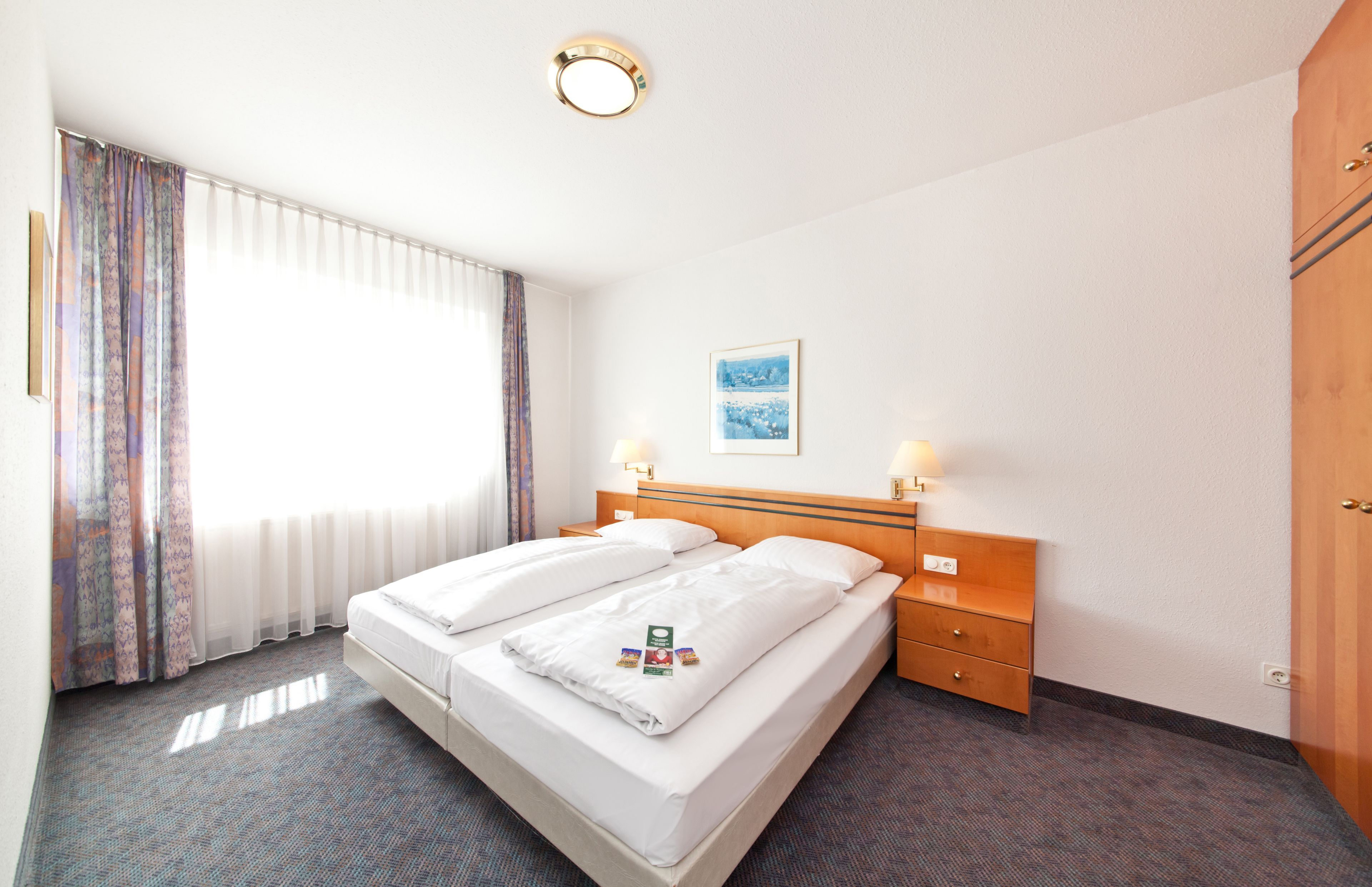 Una habitación de hotel con una cama doble, dos mesitas de noche con lámparas, un armario de madera, un cuadro enmarcado en la pared y una ventana con cortinas de encaje.