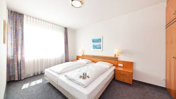 Una habitación de hotel con una cama doble, dos mesitas de noche con lámparas, un armario de madera, un cuadro enmarcado en la pared y una ventana con cortinas de encaje.
