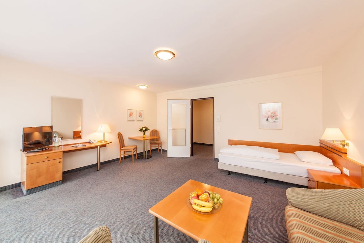 A hotel room with a bed, a desk with a chair, a small table with fruit, a sofa, and a door leading to another room.